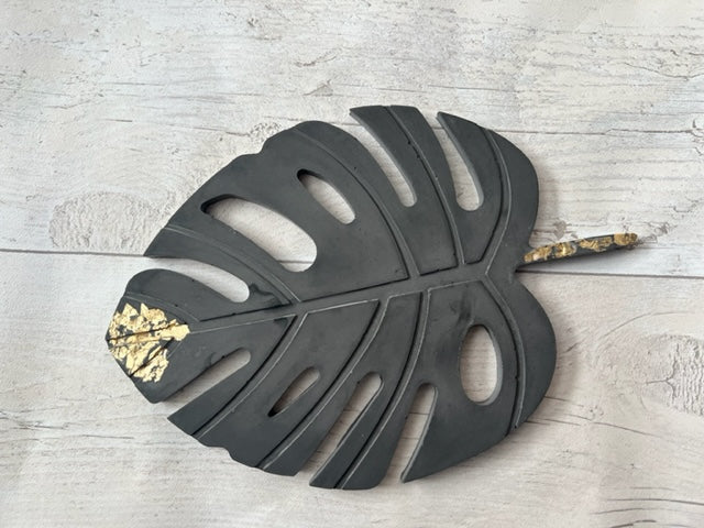 A dark charcoal grey Monstera leaf coaster with gold foil detail, handmade from eco resin.